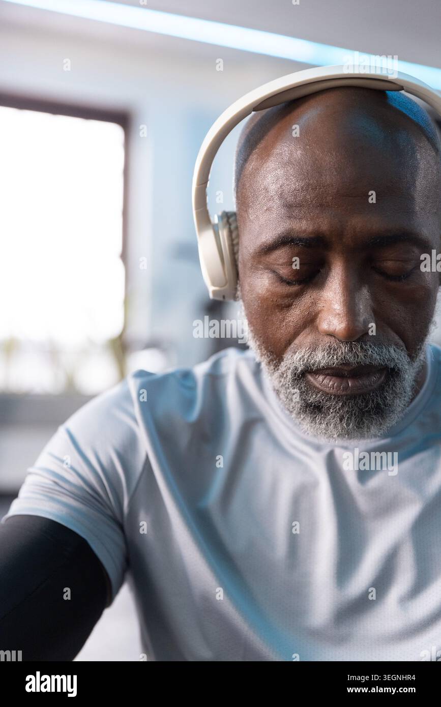 Homme afro-américain senior assis à la salle de gym portant un casque blanc et un manchon de compression noir Banque D'Images