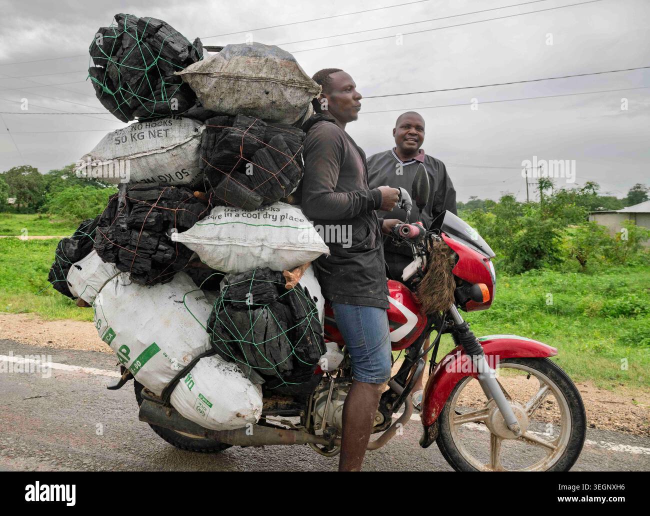 Un homme équilibre une énorme cargaison surchargée de sacs de charbon de bois sur une moto sur une route kenyane comme un compagnon regarde avec amusement. Banque D'Images