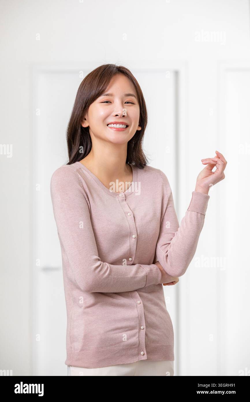 Une jeune femme asiatique se tient debout dans une pièce blanche lumineuse, souriant et faisant des gestes avec sa main. Banque D'Images