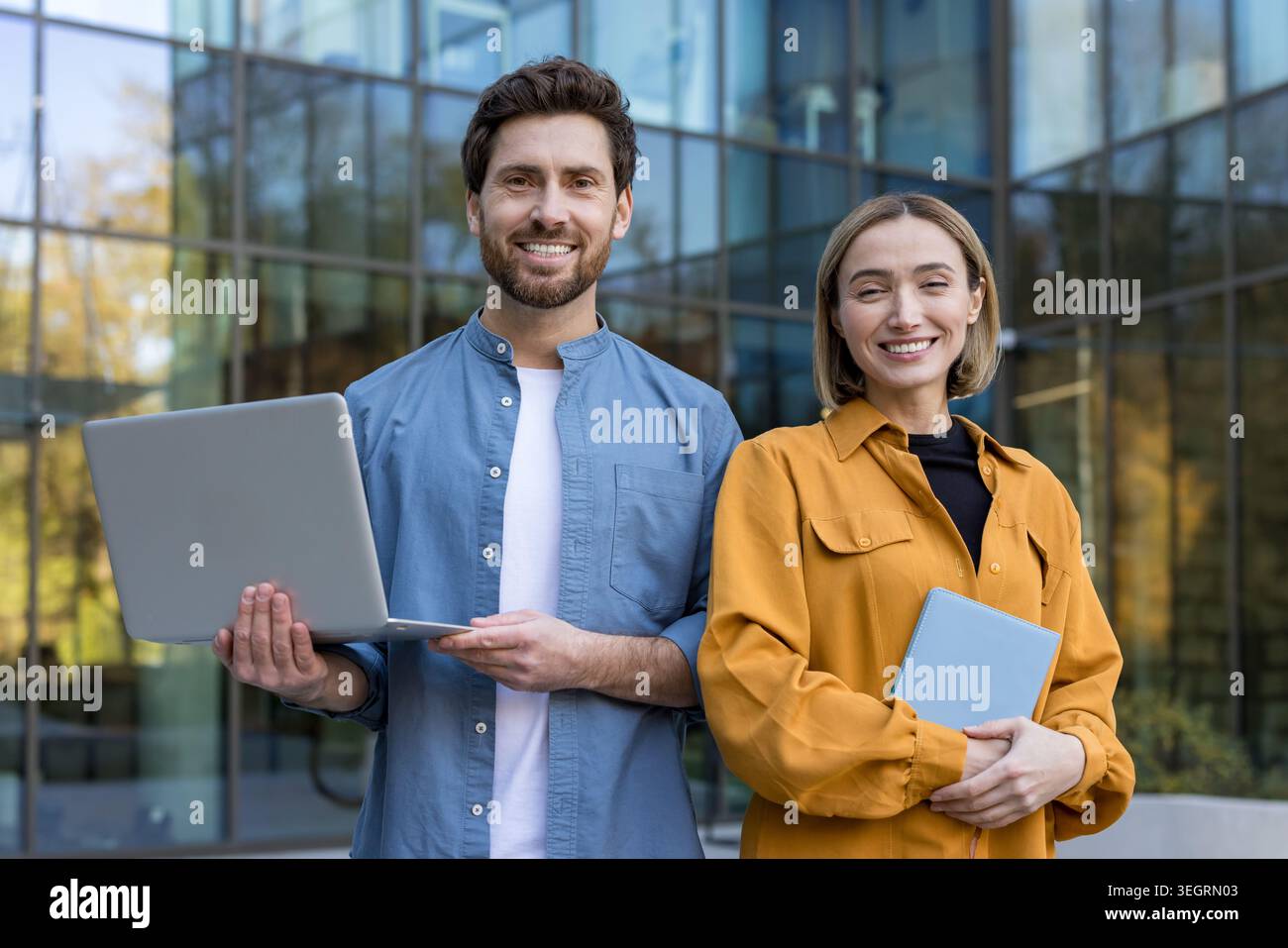 Jeunes collègues d'affaires debout à l'extérieur près d'un immeuble de bureaux en verre moderne, souriant à la caméra tout en partageant un ordinateur portable et une tablette, transmettant le travail d'équipe Banque D'Images