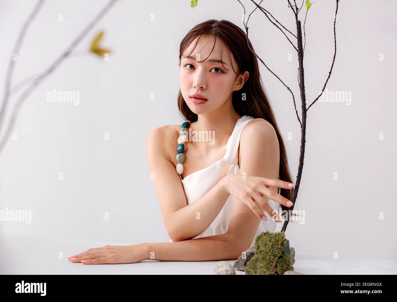 Une jeune femme est assise à une table blanche, sa main reposant sur un rocher recouvert de mousse, entourée d'éléments naturels. Banque D'Images