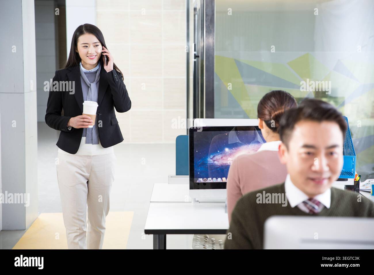 Une jeune femme asiatique en costume d'affaires entre dans un bureau moderne, parlant au téléphone tout en tenant une tasse de café. Banque D'Images