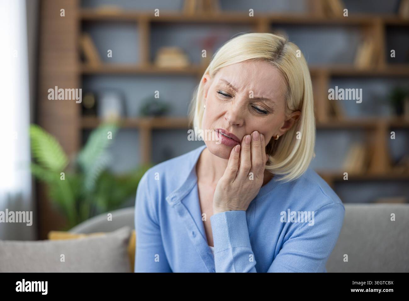Femme blonde mature éprouvant des douleurs dentaires sévères et de l'inflammation, assise à la maison et touchant sa joue, ayant besoin de soins dentaires urgents et de soulagement des problèmes de santé Banque D'Images