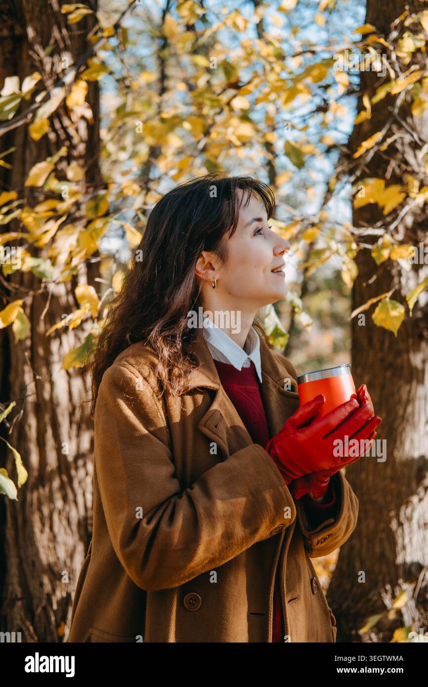 Femme se tient dans la forêt tenant le récipient de boisson tout en regardant vers le haut. Accents de vêtements de couleur audacieuse, style de vêtements d'extérieur haut de gamme commercial, élégant Banque D'Images