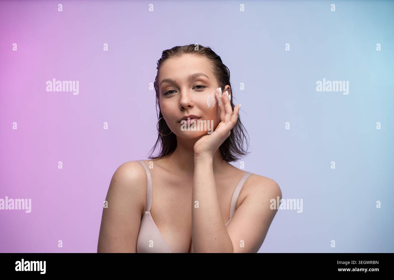 Jeune femme appliquant une crème pour le visage sur sa joue, favorisant une routine de beauté quotidienne pour une peau saine sur un fond coloré avec un espace de copie, en se concentrant sur Banque D'Images