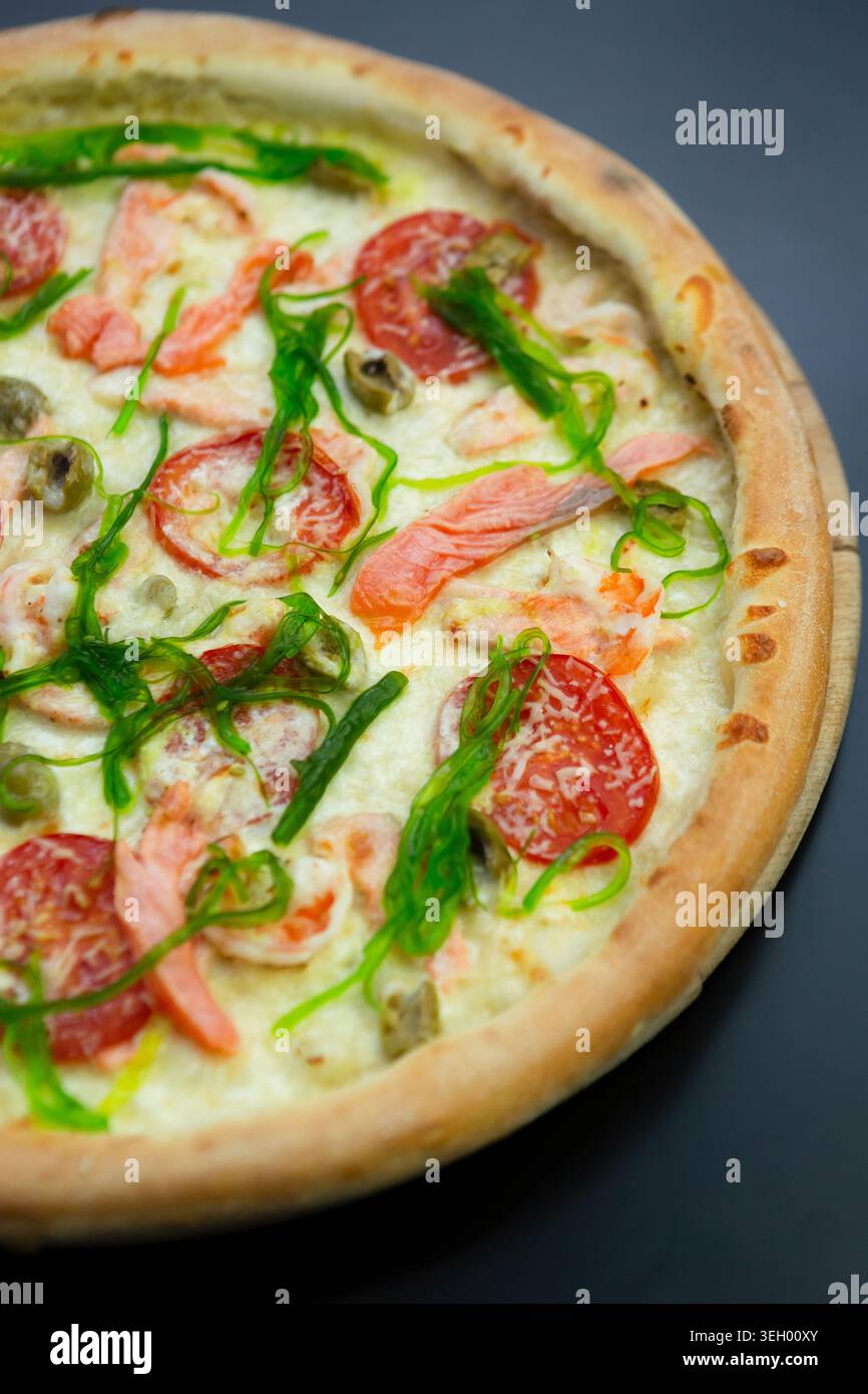 Une pizza est placée sur une planche de bois, surmontée de saumon, tomates, oignons verts et olives. La scène montre une atmosphère chaleureuse adaptée pour profiter de f Banque D'Images