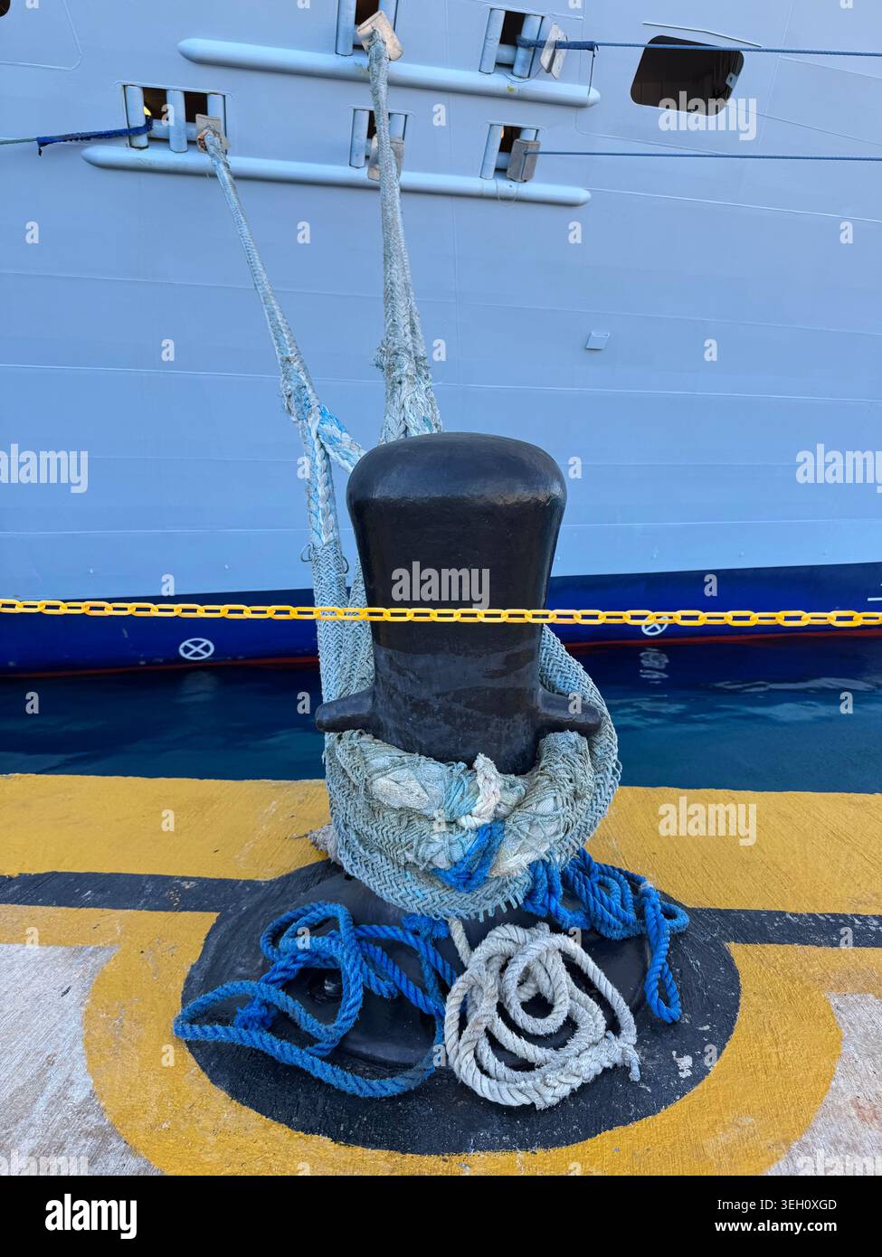 Une lourde borne d'amarrage noire avec des cordes enroulées autour d'elle sur un plancher de jetée peint en jaune et bleu contre un mur de bateau de croisière à Cabo Rojo. Banque D'Images