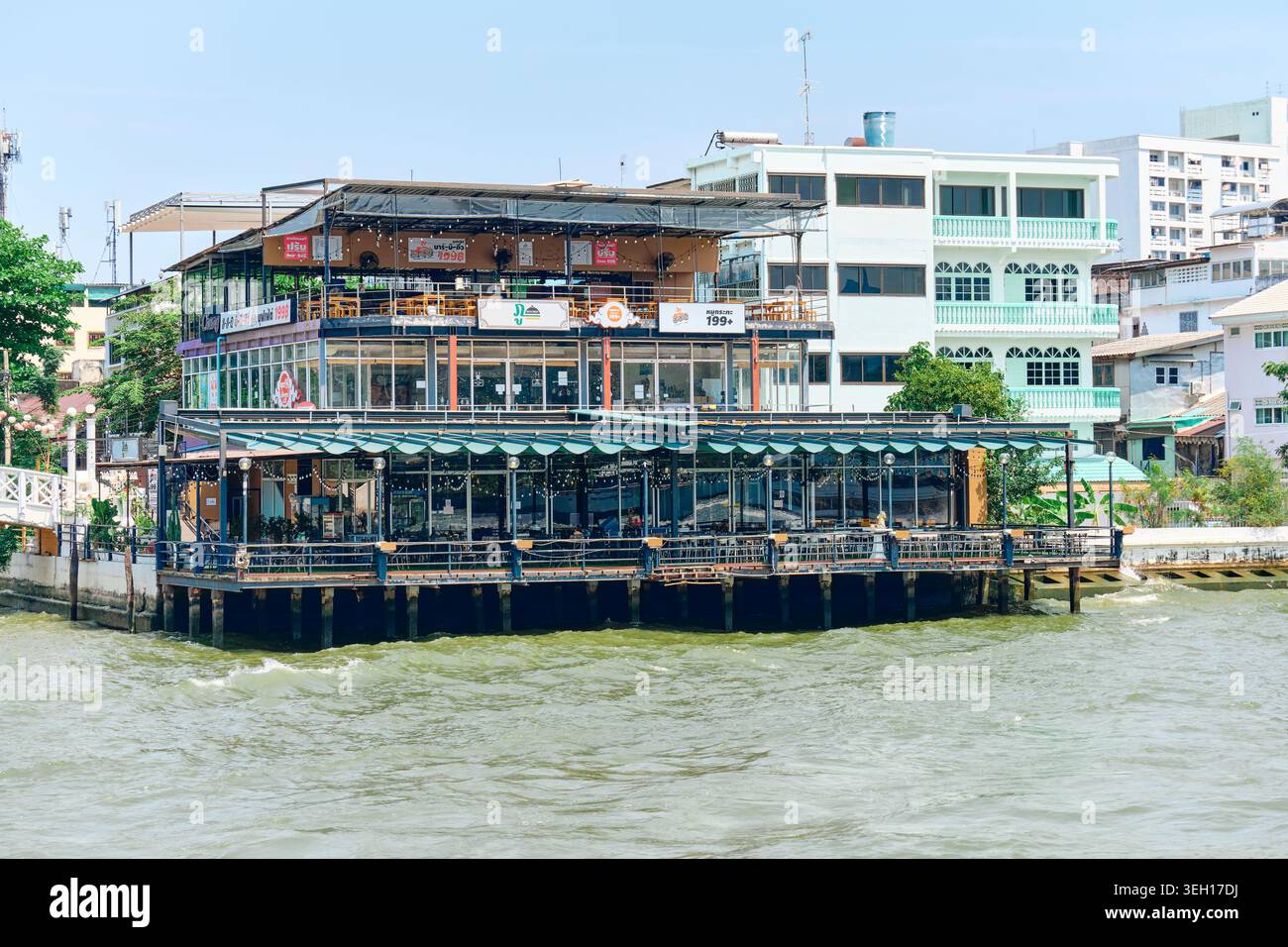 BANGKOK, THAÏLANDE - 27 février 2025 : Café au bord de la rivière sur pilotis au-dessus de la rivière Chao Phraya à Bangkok avec de grandes fenêtres en verre et une terrasse ouverte. Banque D'Images