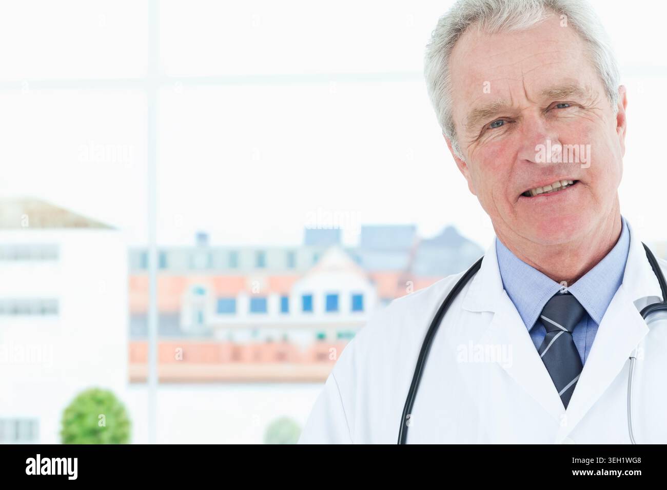 Médecin masculin principal debout à la fenêtre de la clinique portant une blouse blanche de laboratoire et un stéthoscope, espace de copie Banque D'Images