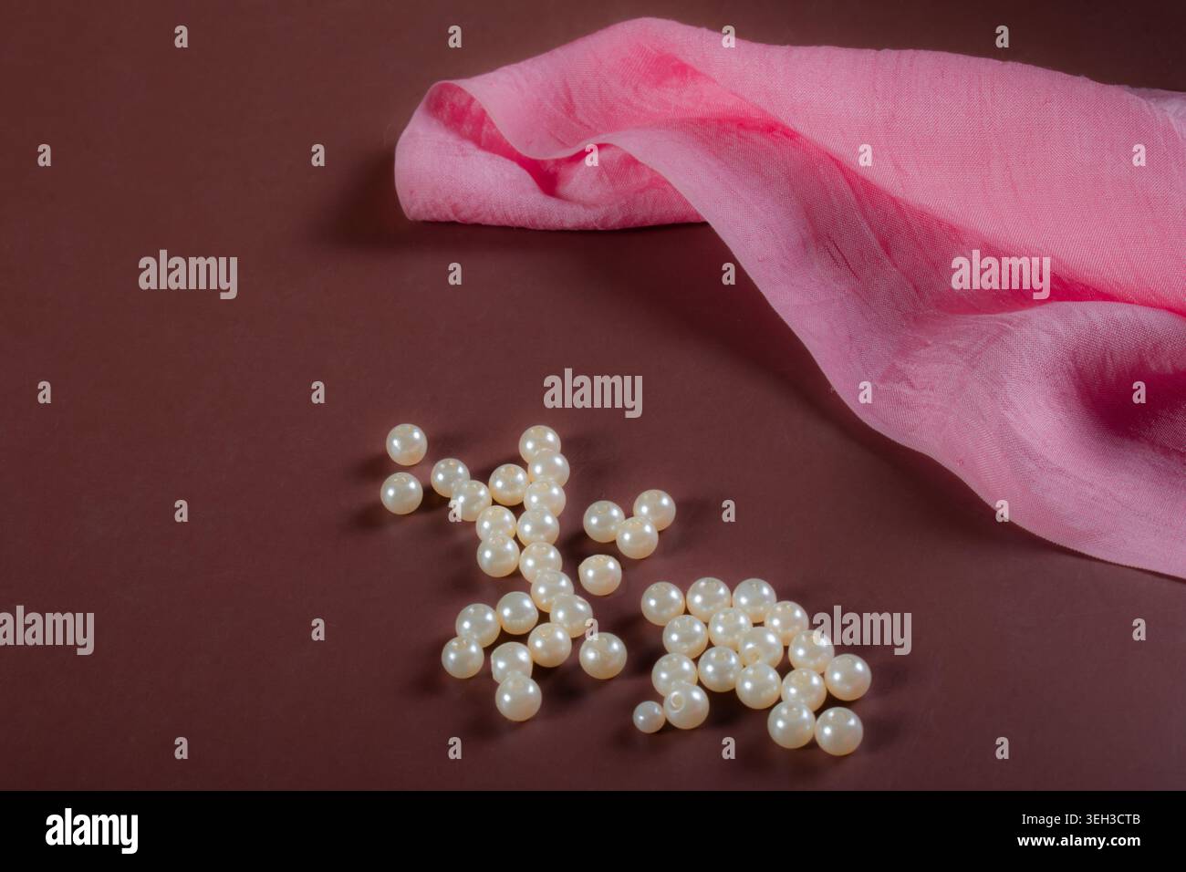 Perles décoratives blanches placées à côté d'un tissu de soie de mûrier rose doux sur une surface marron, créant une élégante composition de nature morte sur le thème de la mode et du textile. Banque D'Images
