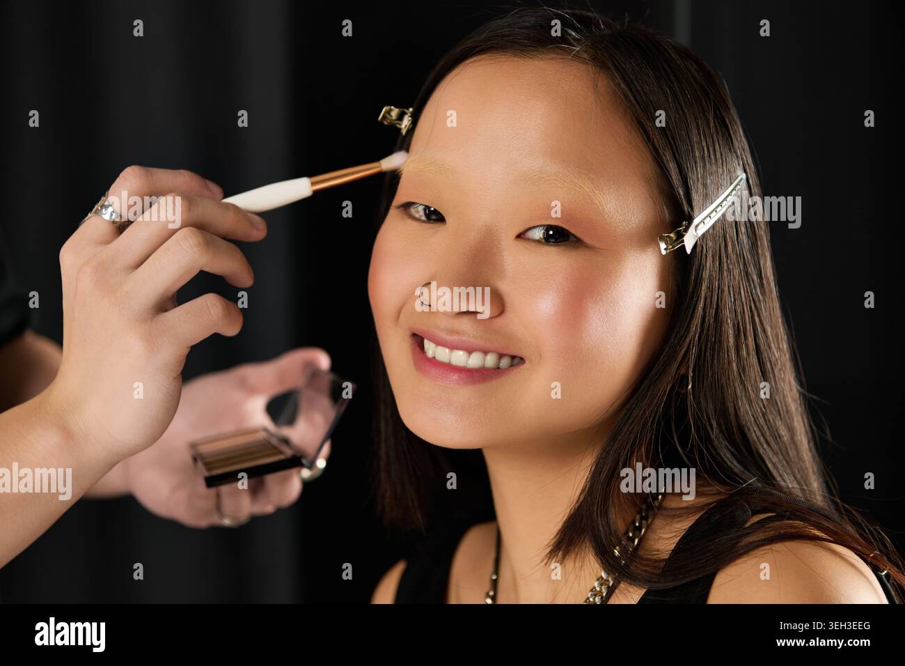 Un modèle sourit comme un maquilleur applique un maquillage impeccable, mettant en valeur l'énergie vibrante des coulisses. Banque D'Images