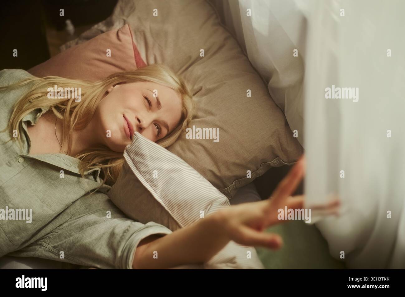 Jeune femme caucasienne adulte se reposant dans son lit, tendant vers le rideau et se réveillant à la maison. Représentant la routine matinale, le bien-être du sommeil, la publicité de style de vie, plan horizontal Banque D'Images