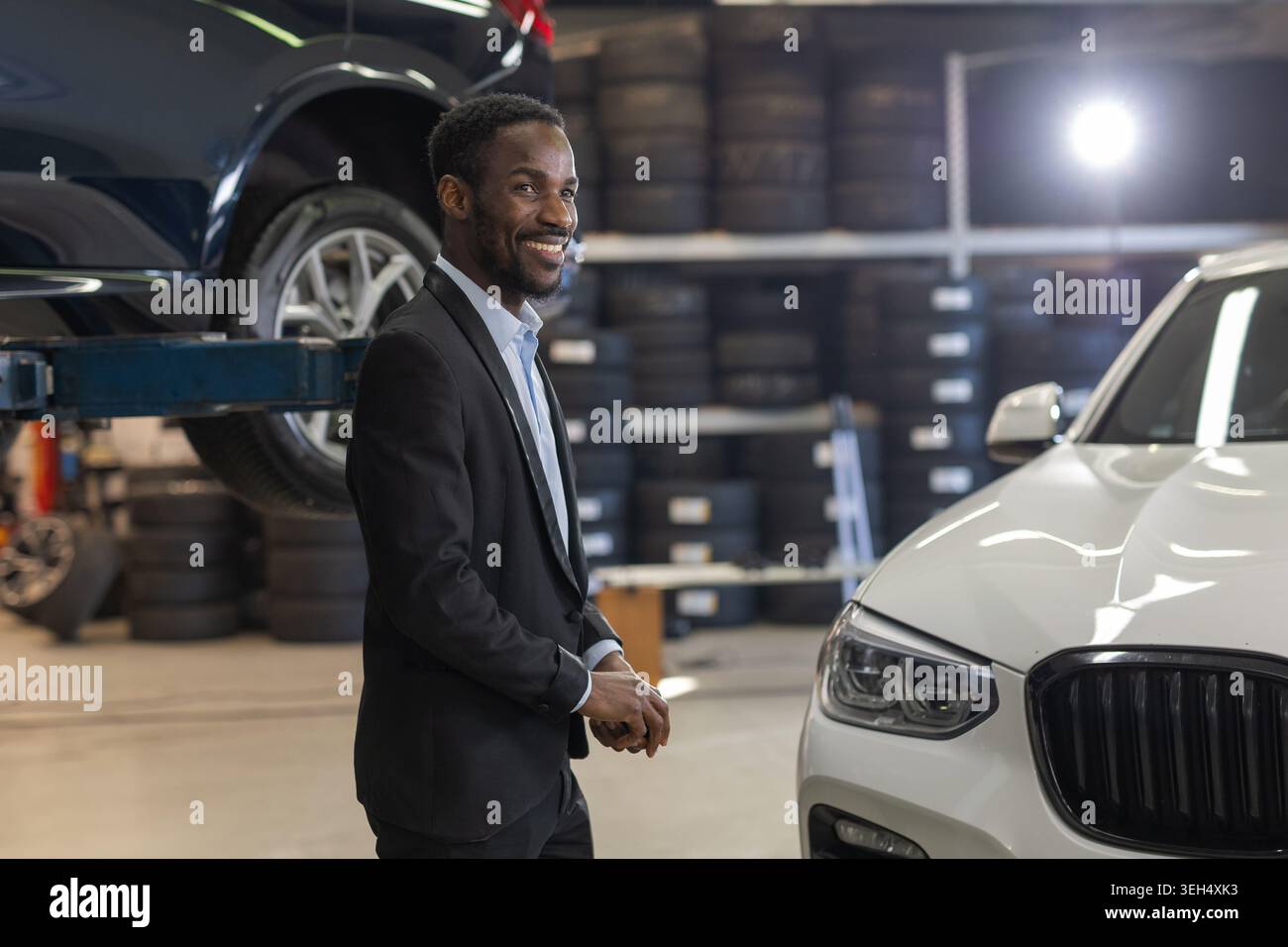 Un homme professionnel se tient dans un atelier de réparation automobile entouré de voitures et de mécaniciens travaillant. Banque D'Images