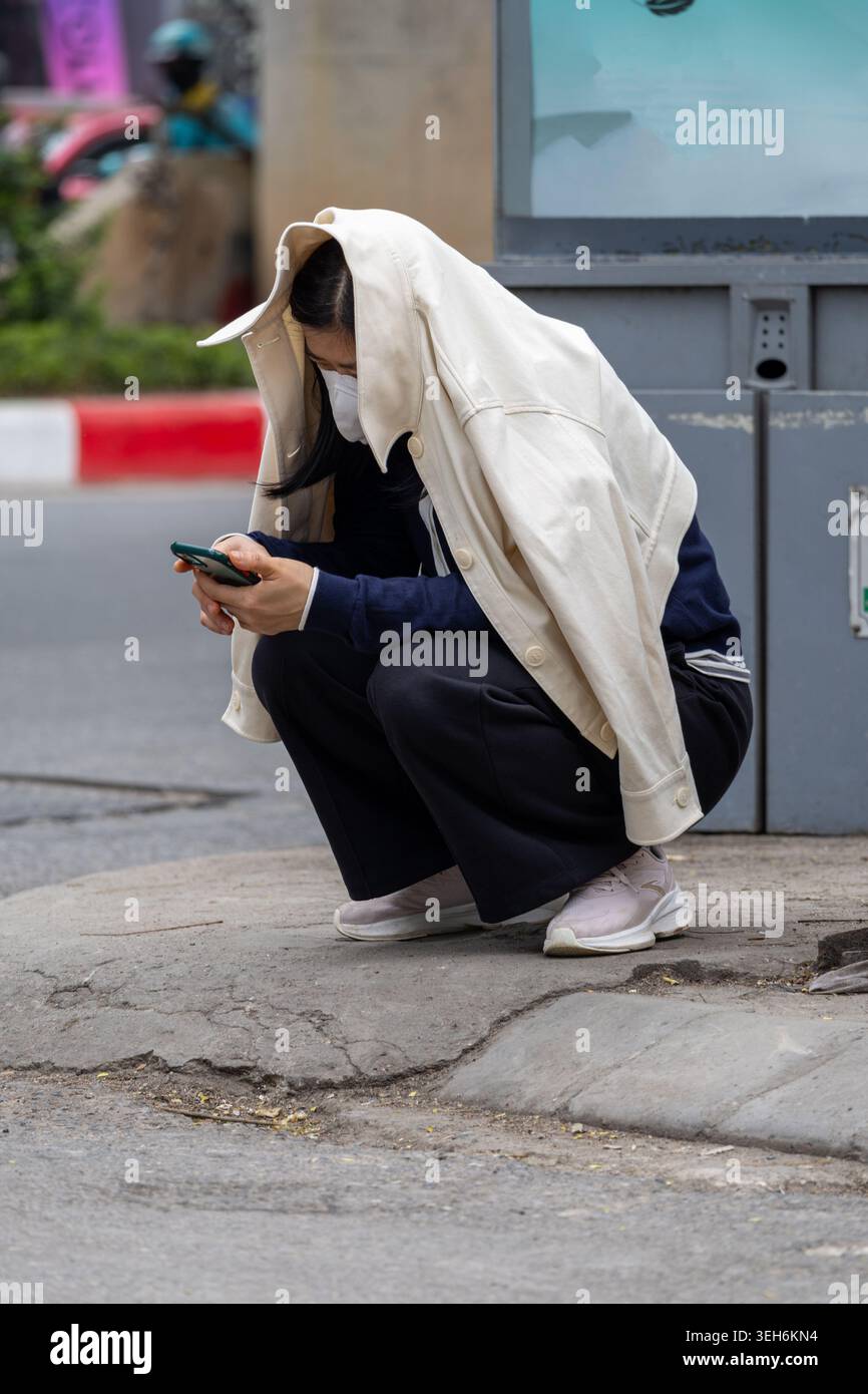 Jeune femme accroupie sous un tissu blanc tout en utilisant smartphone à l'extérieur dans la scène de rue urbaine Banque D'Images