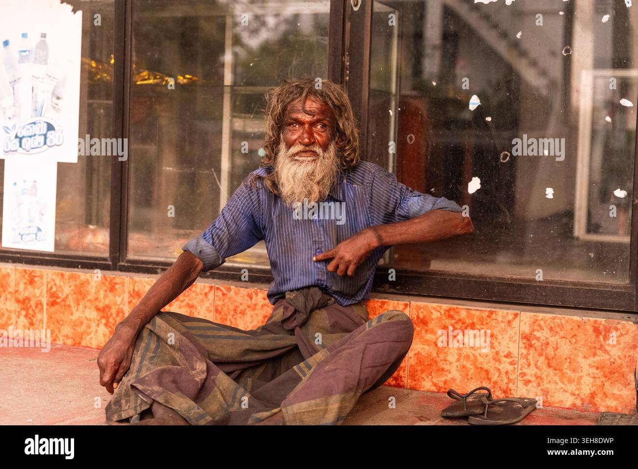 Sri Lanka - 14 janvier 2026 : vue d'un homme âgé aux longs cheveux mats et à la barbe portant une chemise bleue et un sarong assis devant un verre Banque D'Images