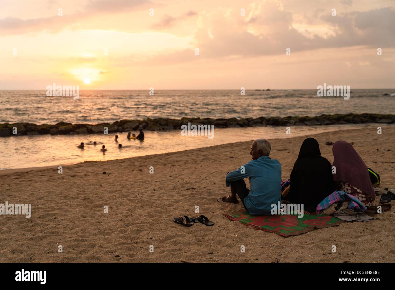Sri Lanka - 14 janvier 2026 : vue de personnes assises sur une plage de sable et d'autres nageant dans l'océan pendant un coucher de soleil doré au ciel nuageux. Banque D'Images