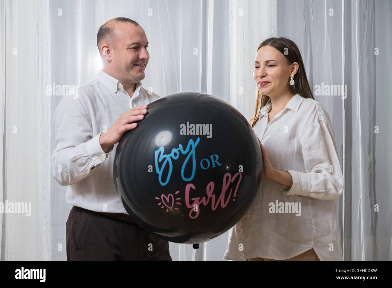 Couple blanc tenant le ballon de fête de genre, grand ballon noir lit garçon ou fille, future mère et père souriant dans des chemises blanches décontractées, debout Banque D'Images