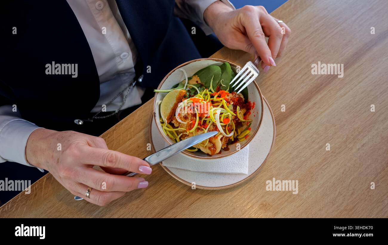 Femme d'affaires senior mangeant de la salade dans le café, légumes vibrants avec fourchette et couteau sur la table en bois, bol en porcelaine, filtre naturel de la lumière du soleil Banque D'Images