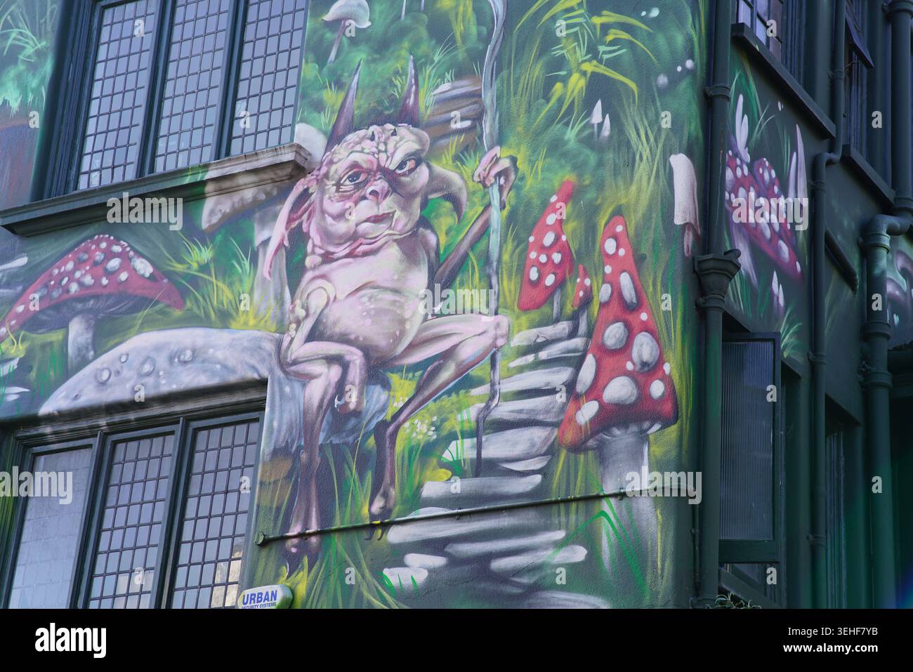 Créature fantaisiste fantaisiste ressemblant à un gobelin assis sur des marches de pierre entourée de champignons géants à taches rouges dans une rue magique et verdoyante de la forêt Brighton Banque D'Images