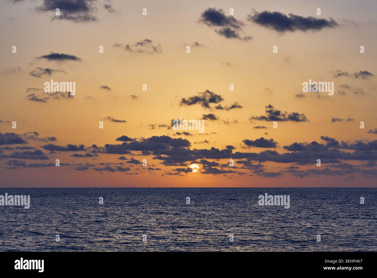 Soleil plongeant sous l'horizon, peignant le ciel avec des couleurs chaudes et des nuages dispersés sur l'océan calme Banque D'Images