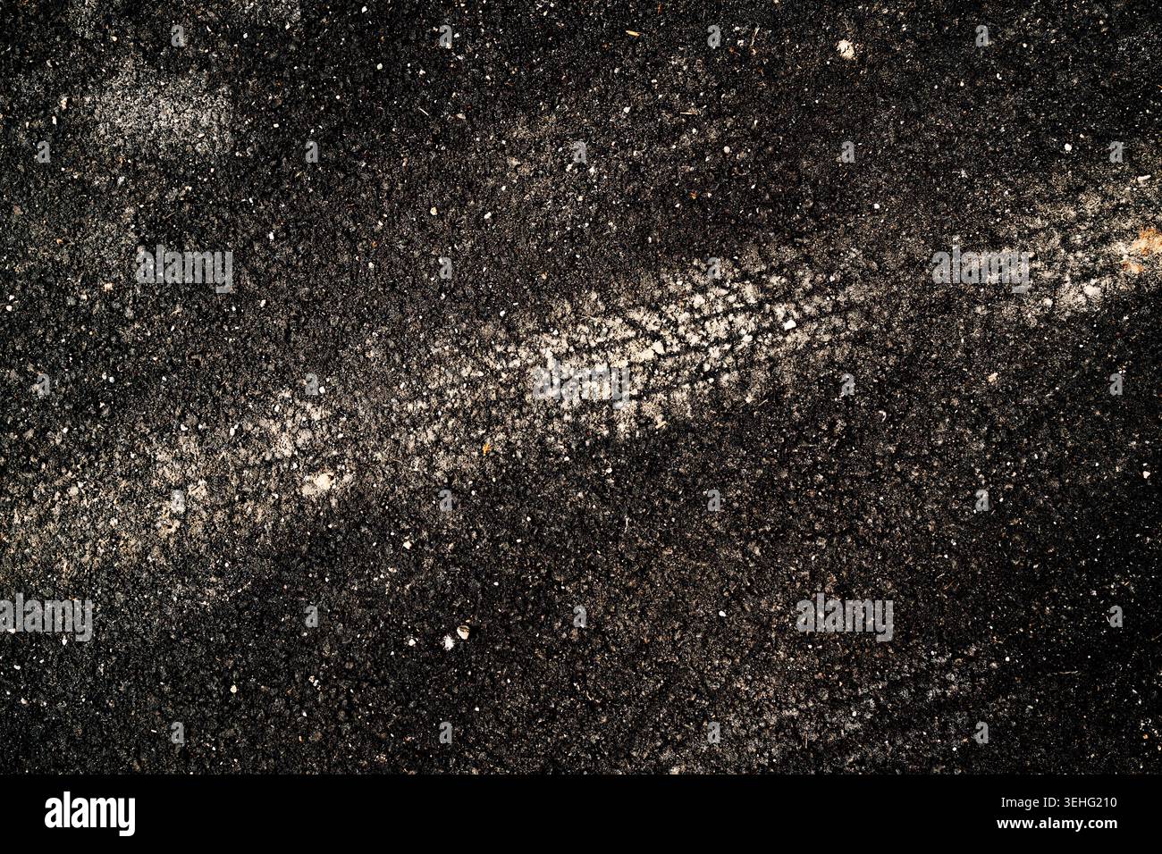 Gros plan d'une voie à pneu unique enfoncée dans une chaussée asphaltée sombre, mettant en évidence le motif de la bande de roulement, la texture granuleuse et le contraste. Vue de dessus. Banque D'Images