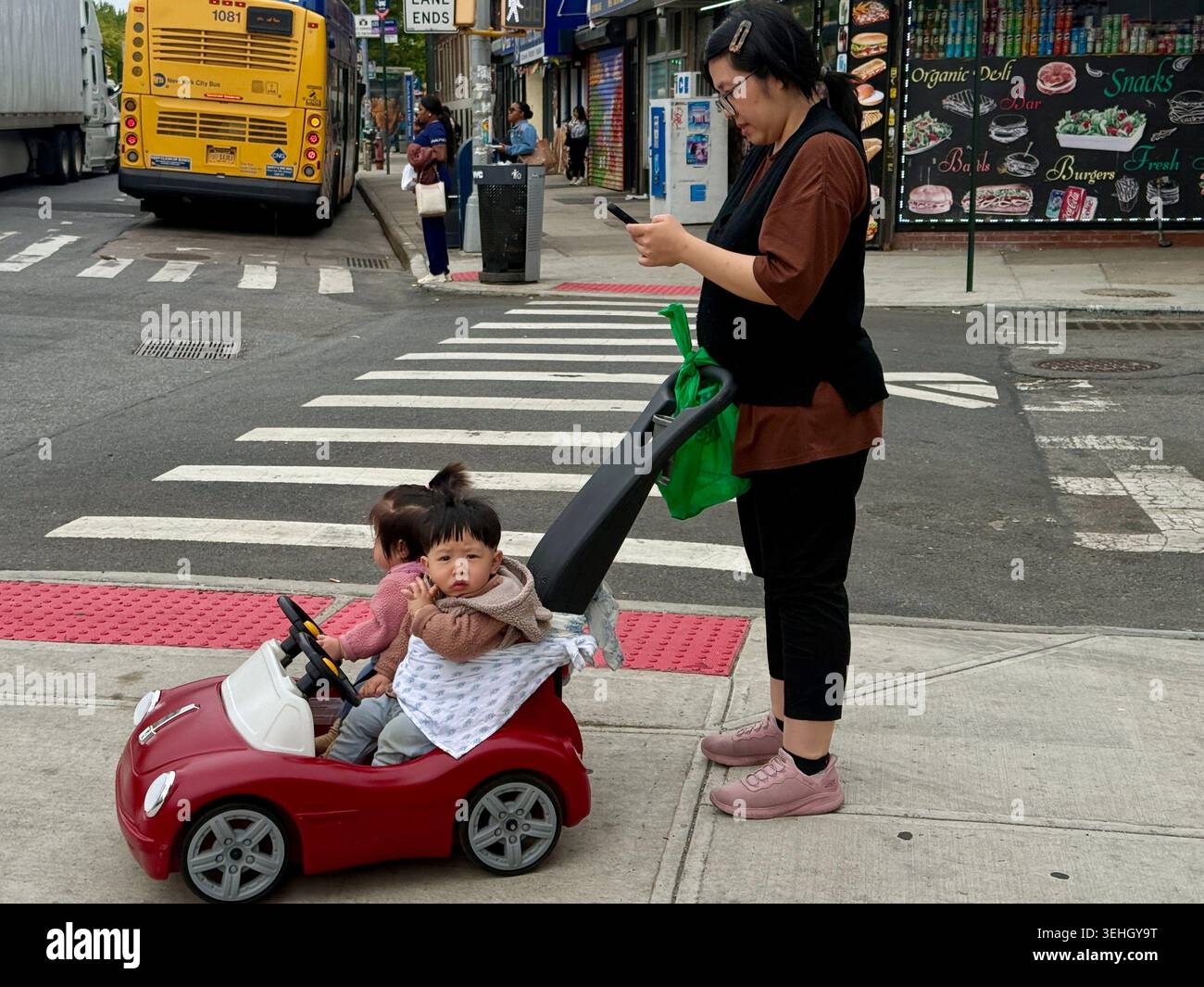 Maman asiatique sur son téléphone avec les enfants dans une poussette de type automobile attendant que le feu de circulation change à Brooklyn, Banque D'Images