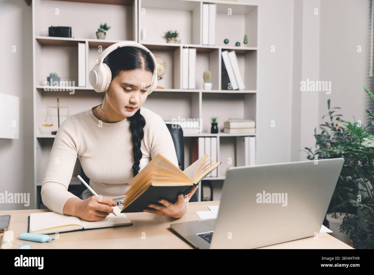 Jeune femme asiatique étudiant à la maison avec des écouteurs, lisant un livre, prenant des notes et utilisant un ordinateur portable au bureau. Apprentissage en ligne, éducation ciblée, à distance Banque D'Images