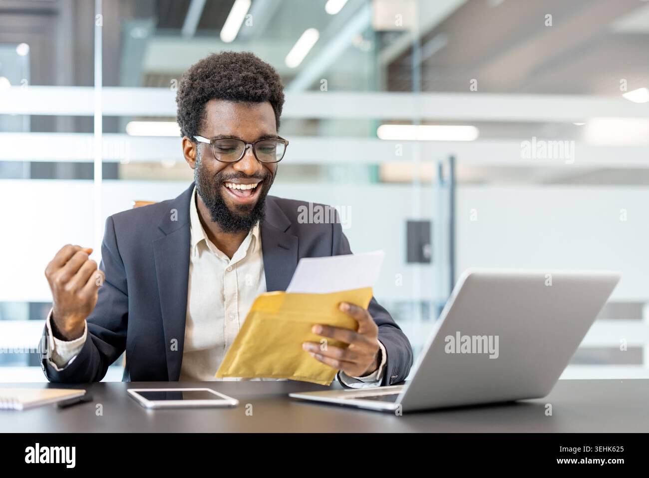 Jeune homme professionnel lisant des nouvelles triomphales à partir d'une enveloppe, se sentant excité et célébrant un résultat réussi ou une promotion dans un espace de travail moderne Banque D'Images