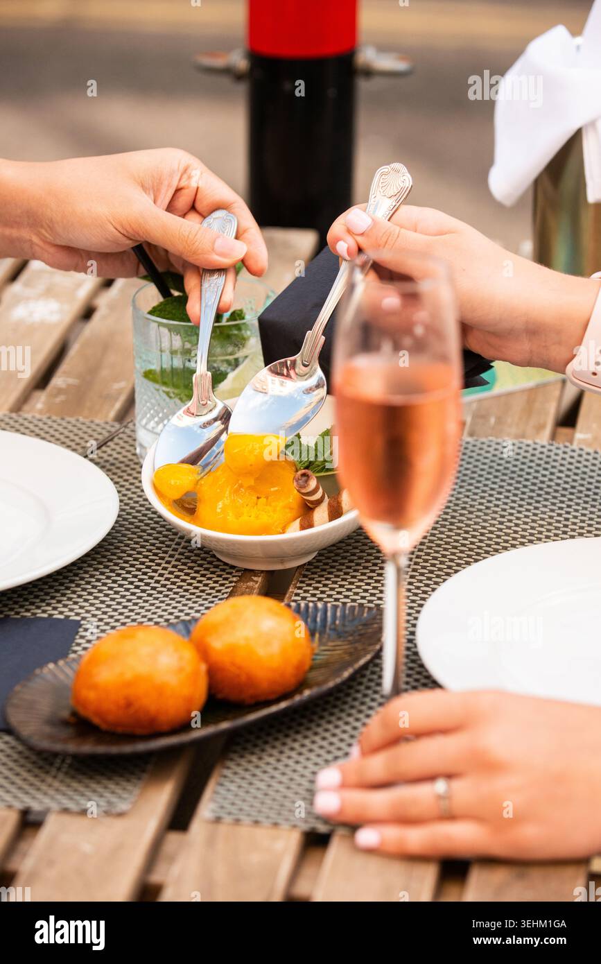 Un gros plan de deux personnes utilisant des cuillères pour partager un bol de sorbet à la mangue sur une table en bois extérieure, avec des cocktails et des petites pâtisseries dans le Forward Banque D'Images