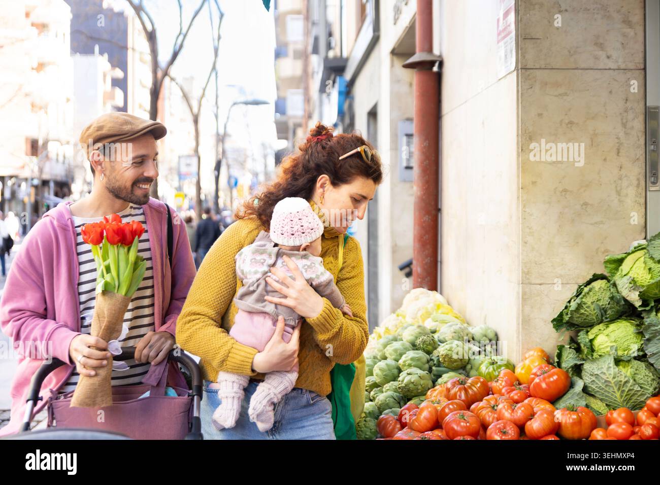 La nouvelle maman souriante tient son nouveau-né tandis que papa porte des fleurs et pousse une poussette, profitant d'un doux moment en famille tout en magasinant des produits frais à Banque D'Images