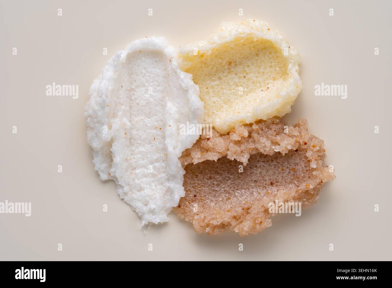 Gros plan de trois frottis de gommage corporel montrant une texture exfoliante crémeuse et granuleuse en blanc, jaune pâle et brun, disposés sur un fond beige doux Banque D'Images