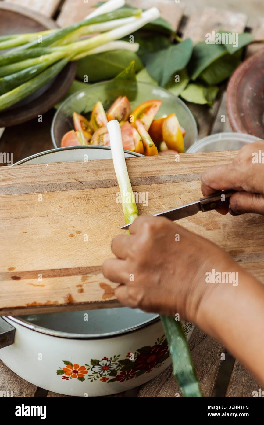 Gros plan de mains coupant de la citronnelle sur une planche à découper en bois, entourées de feuilles de citron vert kaffir et d'épices, capturant la cuisine traditionnelle indonésienne Banque D'Images