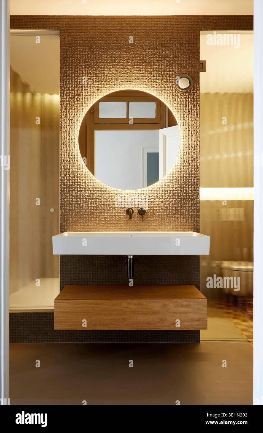 Intérieur de salle de bains contemporain avec meuble-lavabo flottant, miroir rond rétroéclairé, éclairage chaleureux et finitions murales texturées, idéal pour la mise en scène d'une maison Banque D'Images