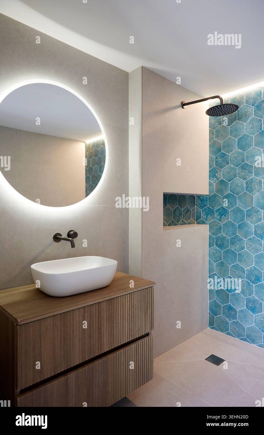 Intérieur contemporain de la salle de bains avec miroir rond rétroéclairé, évier sur meuble-lavabo en bois, robinet noir mat et douche de plain-pied avec carreaux hexagonaux bleus Banque D'Images