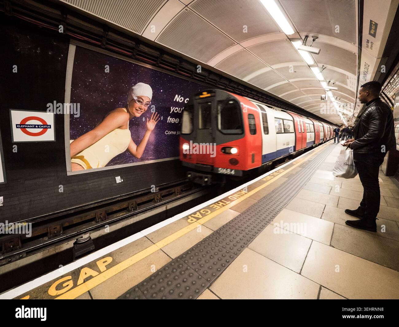 Un train de métro londonien arrive à une plate-forme profonde alors qu'un passager seul attend à côté de l'emblématique bande d'avertissement jaune « Mind the Gap ». Banque D'Images