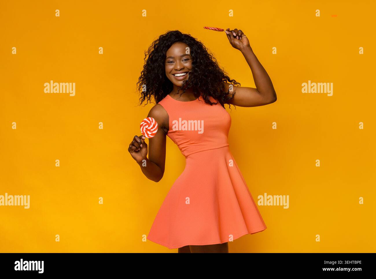 Femme en robe orange tenant joyeusement des sucettes sur fond jaune Banque D'Images