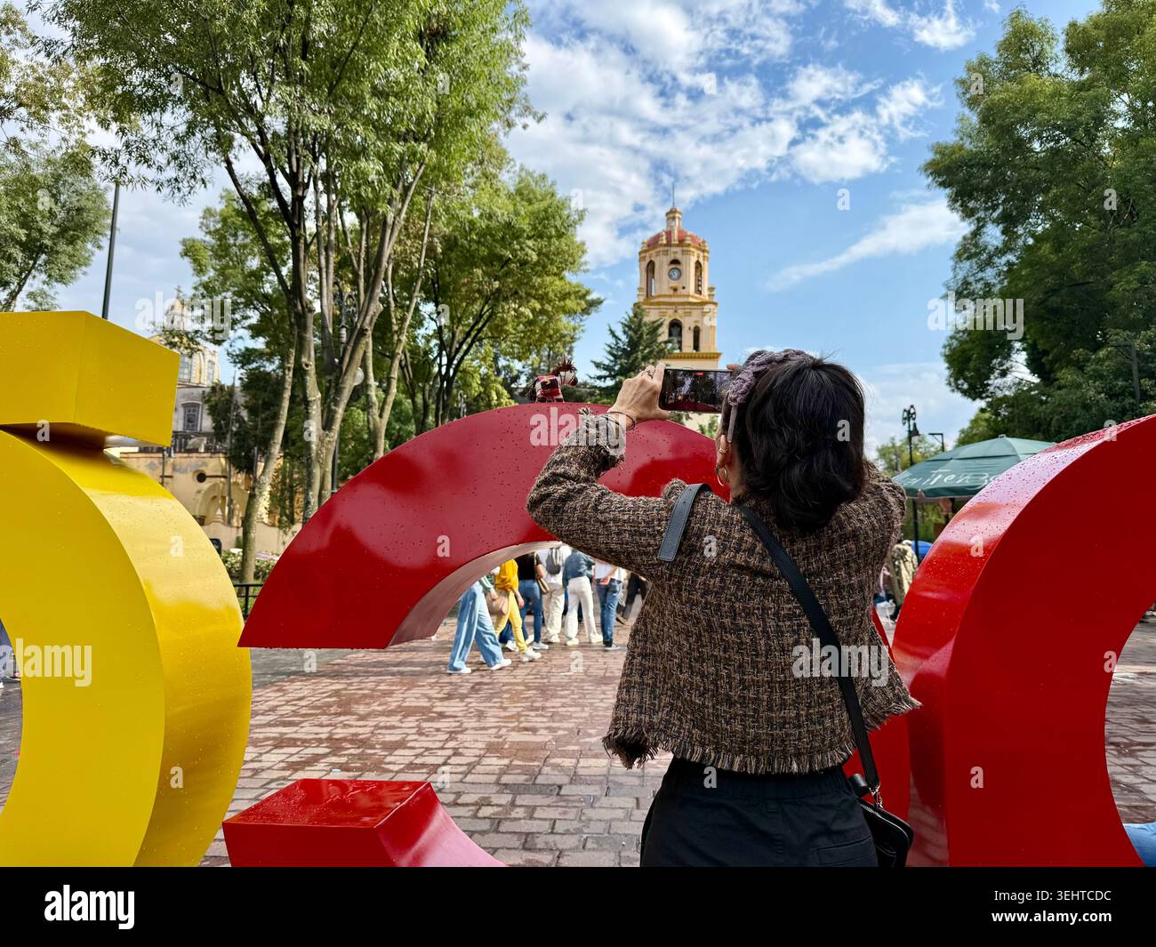 Un visiteur photographie le panneau coloré Coyoacán avec un téléphone portable, avec le clocher de la Parroquia de San Juan Bautista en arrière-plan, Mex Banque D'Images