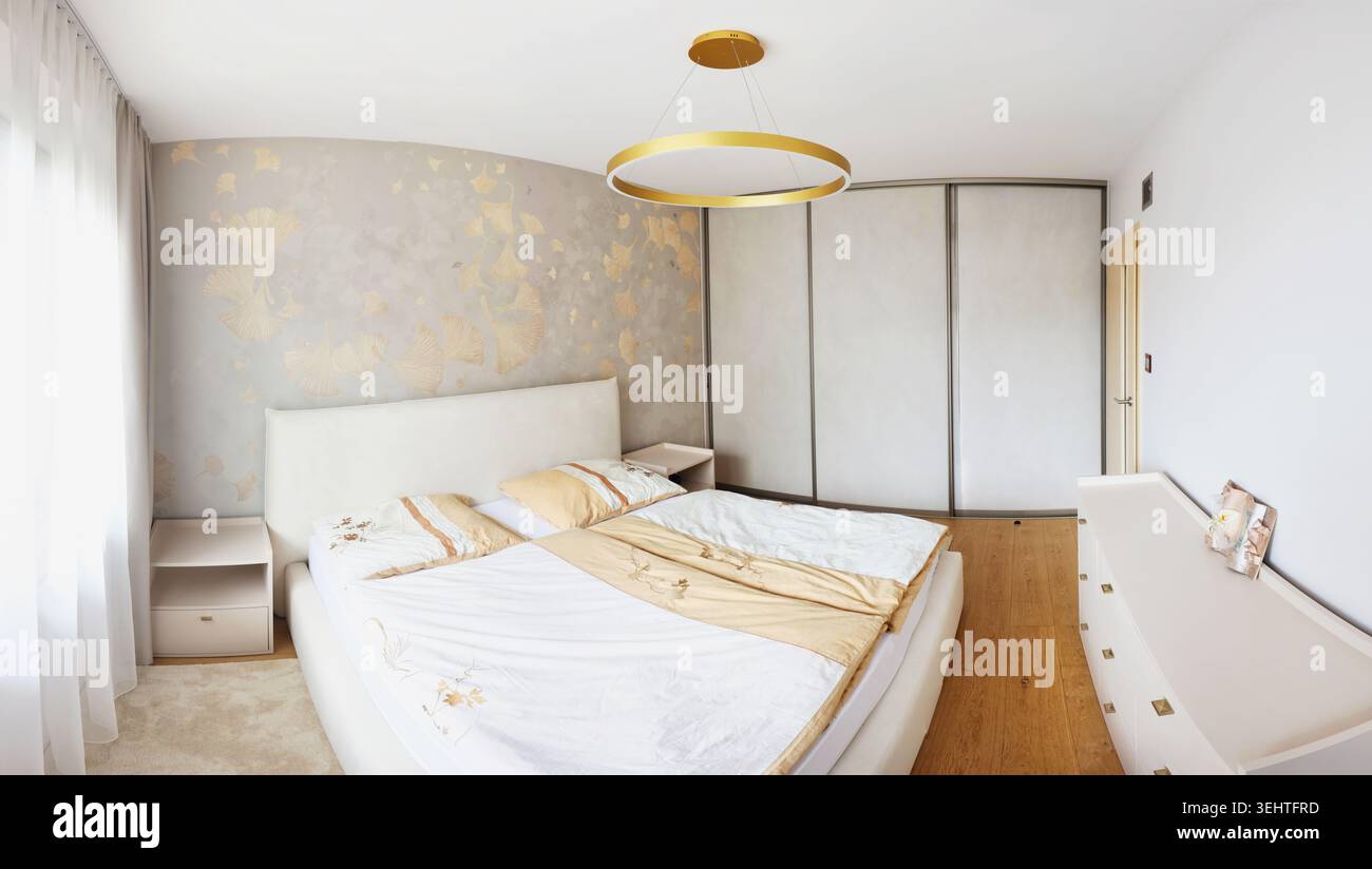 Un intérieur de chambre contemporain avec boiseries, lit beige, fauteuil, suspensions, rideaux doux, et parquet à chevrons. Banque D'Images