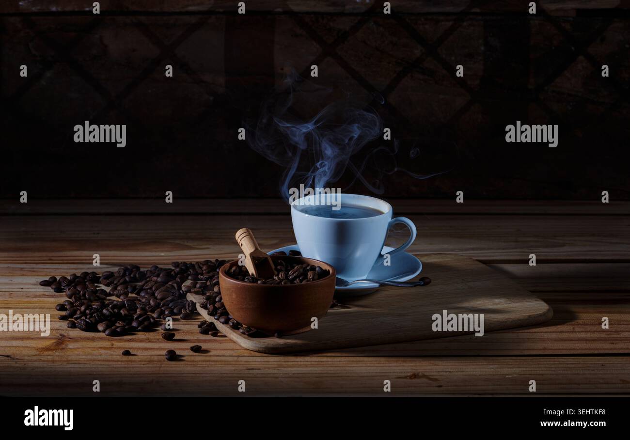 Moody Still Life de café chaud fraîchement infusé dans une tasse en céramique blanche avec de la vapeur visible, placée à côté d'un bol en bois de grains de café torréfiés. Banque D'Images