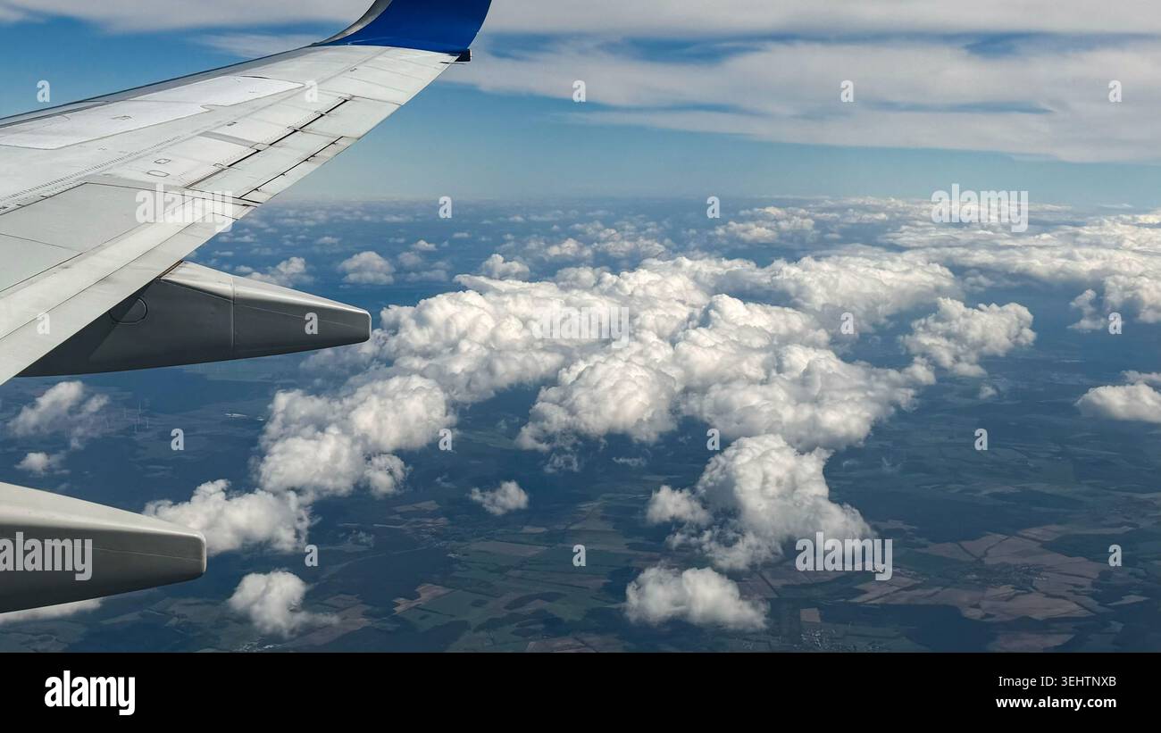 Aile d'avion s'étendant sur des nuages blancs moelleux avec un ciel bleu clair et des champs verts visibles ci-dessous, capturant une vue aérienne sereine d'un vol Banque D'Images