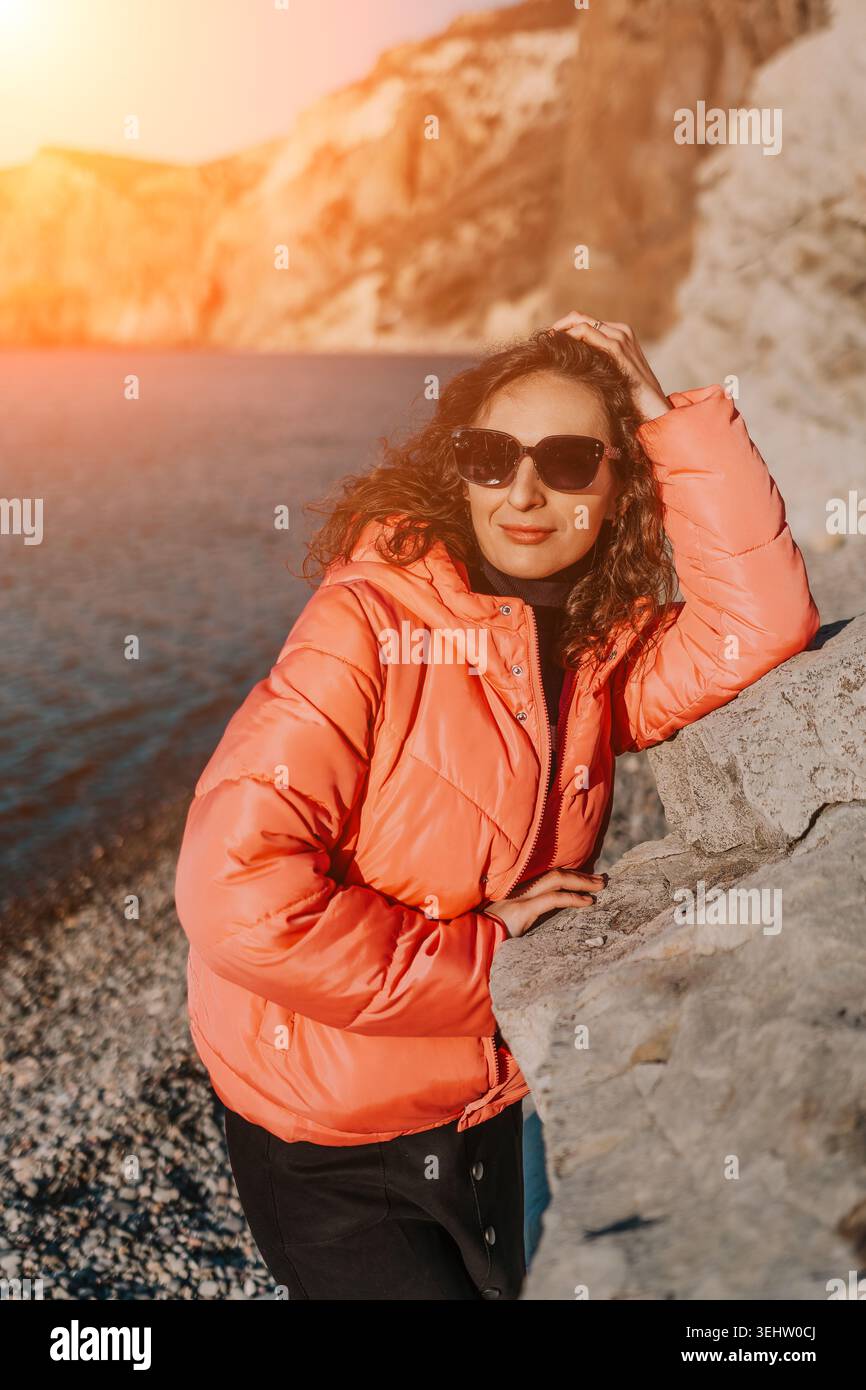 Veste de gonflement de femme côte posant par la mer au coucher du soleil portant des lunettes de soleil sur une plage rocheuse avec des montagnes en arrière-plan Banque D'Images