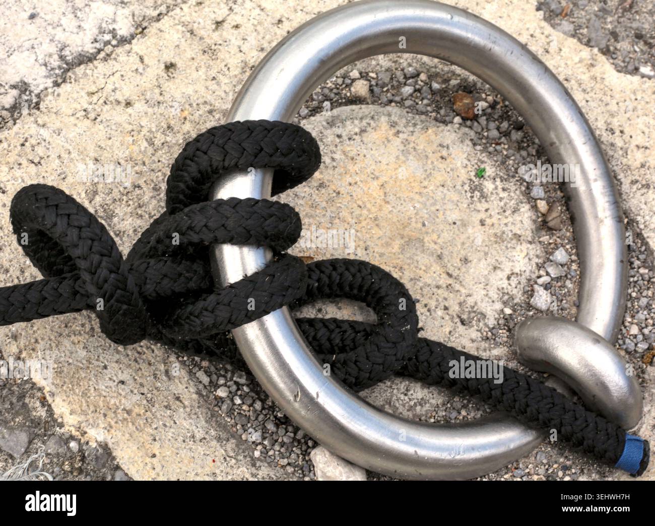 Lourde corde d'amarrage noire nouée solidement autour d'un anneau en acier rouillé sur un quai de jetée en béton. Banque D'Images