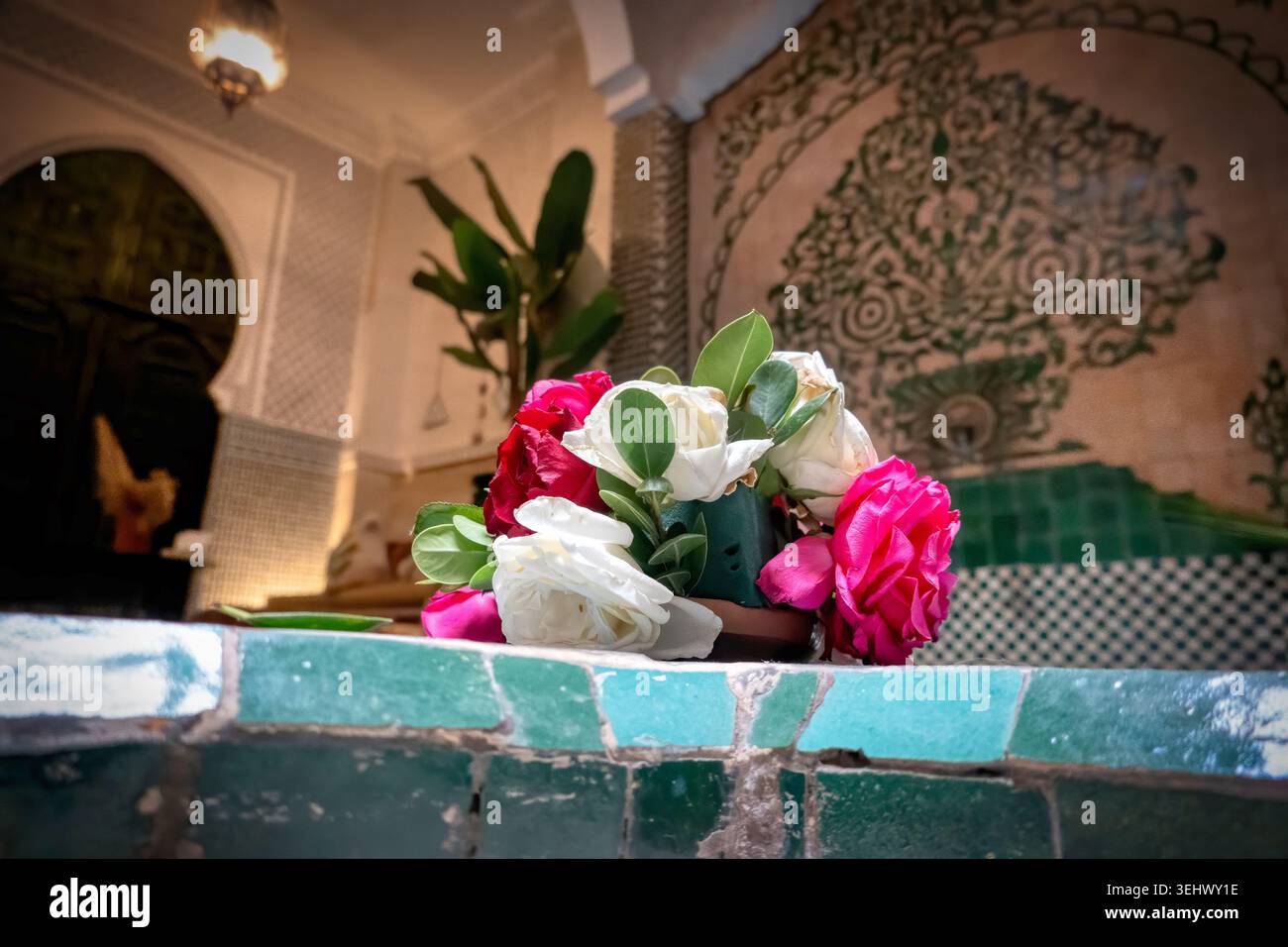 Un bouquet frais de roses roses et blanches repose sur une fontaine carrelée verte dans une cour riad marocaine traditionnelle. Banque D'Images