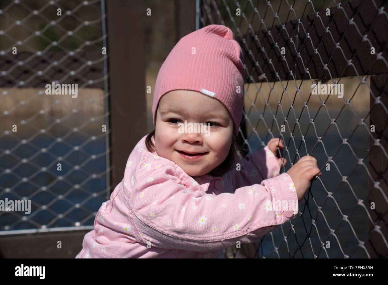 petite fille portant un bonnet rose et veste florale souriant tout en saisissant une clôture en métal à maillons de chaîne sur une jetée en bois ensoleillée par l'eau bleue calme, chaude d Banque D'Images