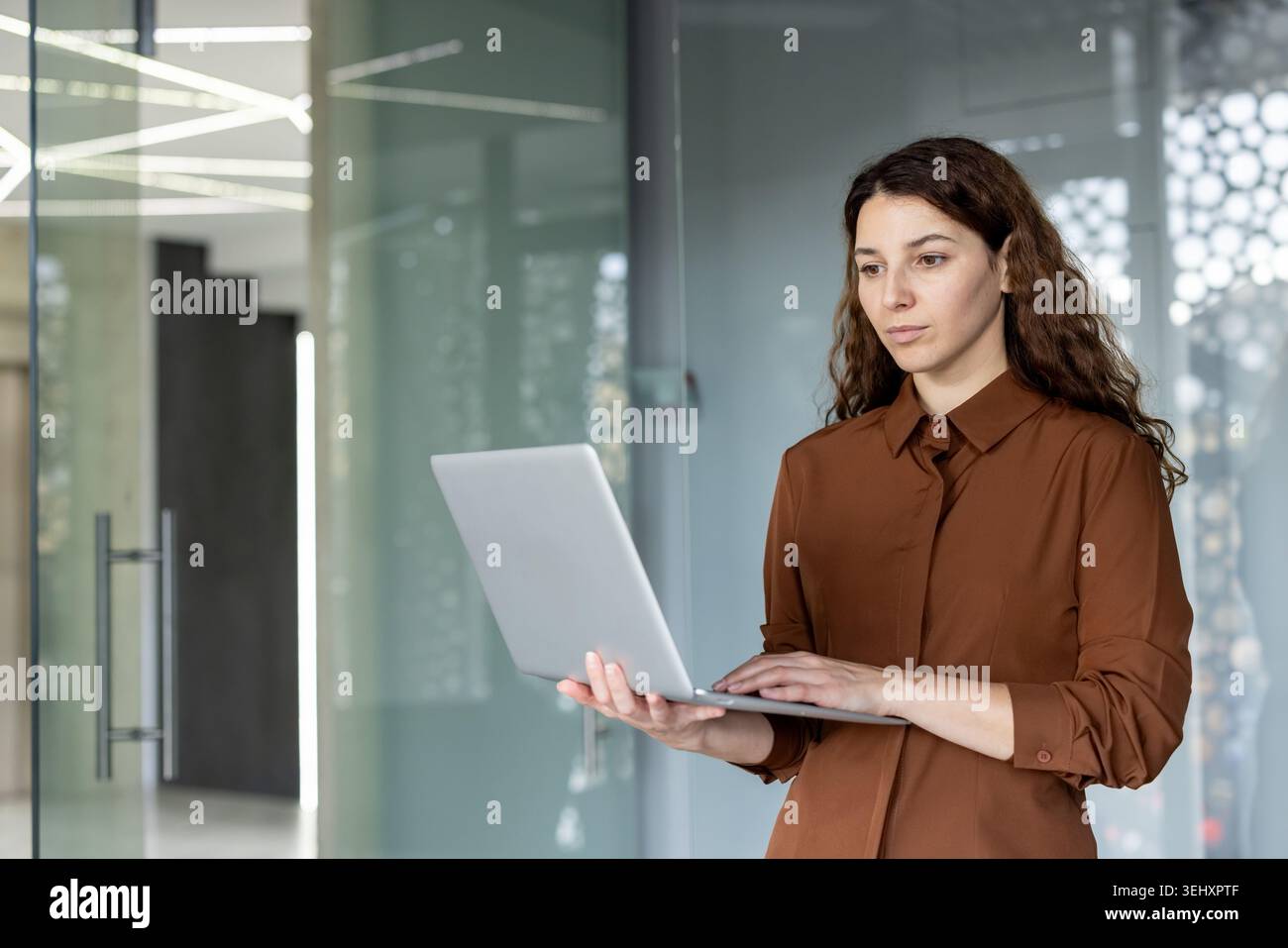 Femme d'affaires professionnelle debout dans un cadre de bureau contemporain, axée sur la saisie sur son ordinateur portable, représentant la flexibilité de travail moderne et l'intégration de la technologie Banque D'Images
