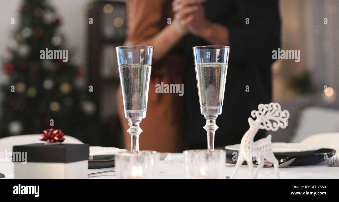 Champagne, noël et cadeau dans la salle à manger de la maison avec couple dansant pour la tradition festive. Alcool, verres et présent sur table dans l'appartement Banque D'Images