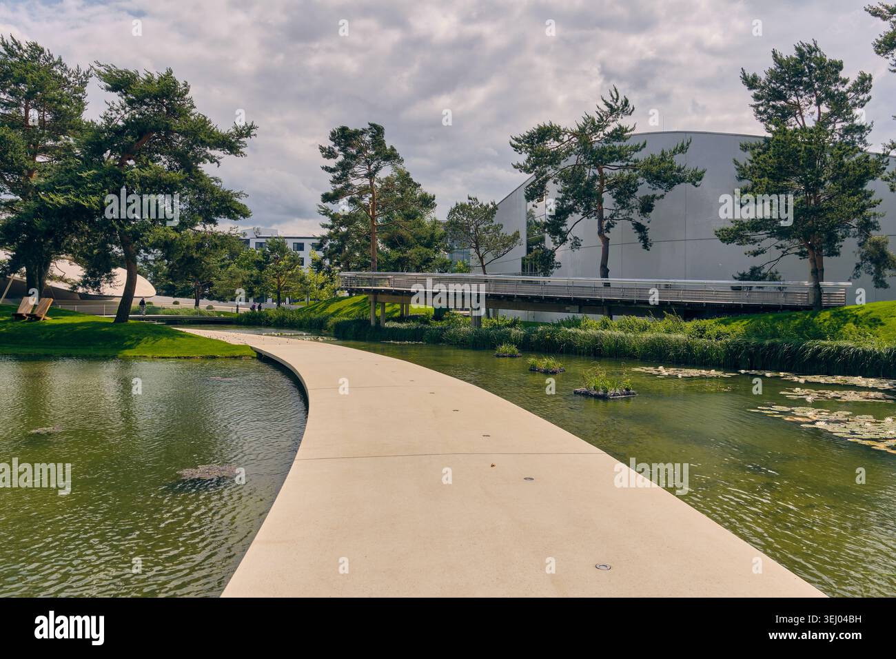 Wolfsburg, Allemagne - 06 juin 2025 : vue sur le parc Autostadt avec une passerelle incurvée au-dessus d'un étang avec des nénuphars, des arbres verts et une arche moderne Banque D'Images