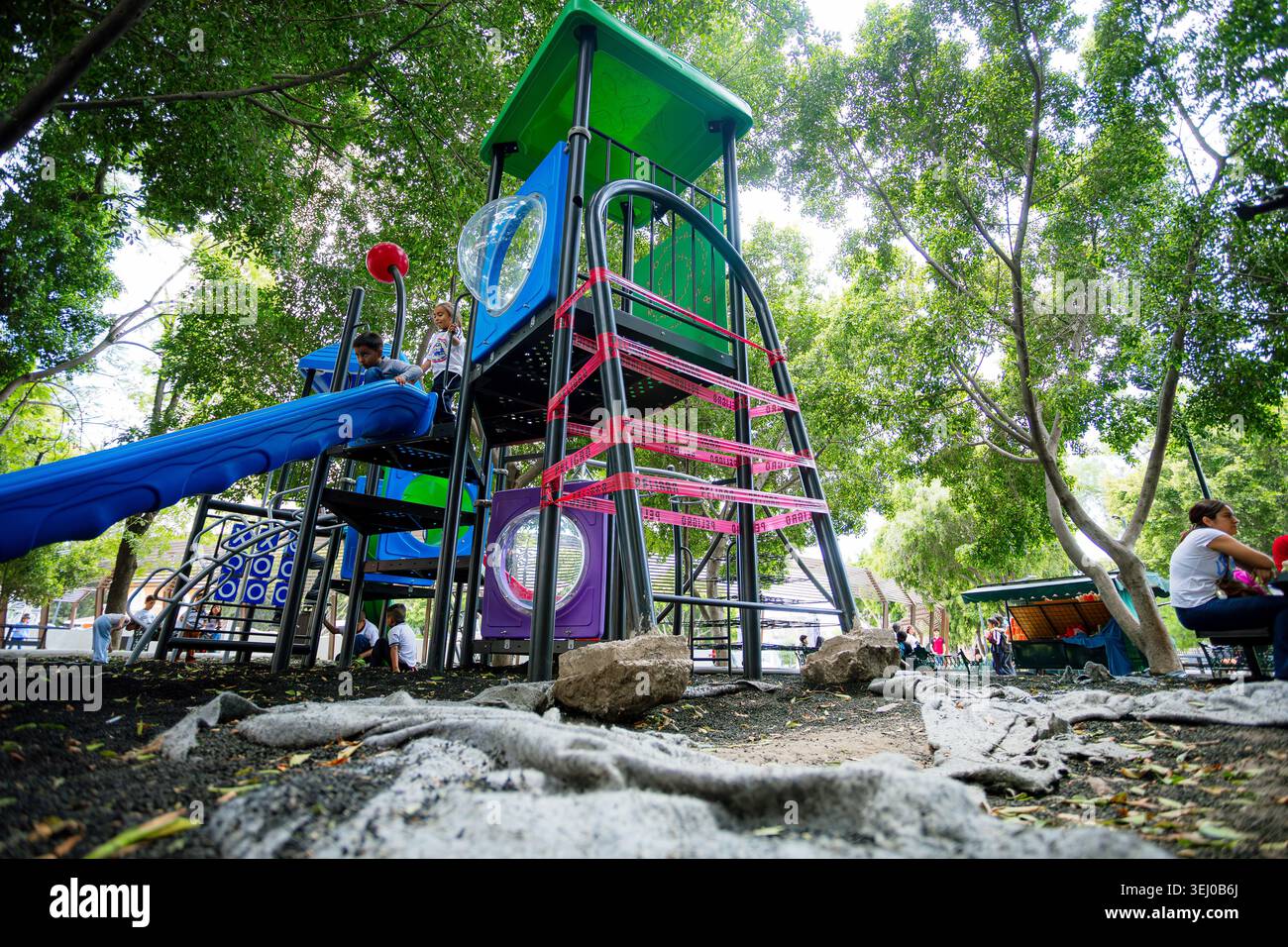 Plusieurs enfants jouent sur une structure colorée de terrain de jeux avec un toboggan bleu, entouré d'arbres verdoyants dans un parc public. Banque D'Images