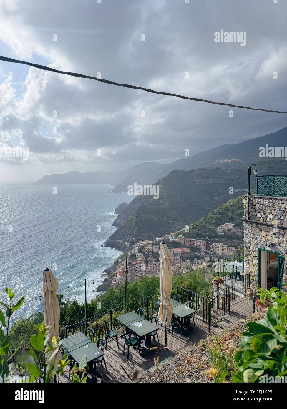 Superbe vue panoramique depuis une terrasse de montagne surplombant un village pittoresque au bord de la mer. Les tables vides avec parasols offrent un cadre serein. Nuages et Banque D'Images