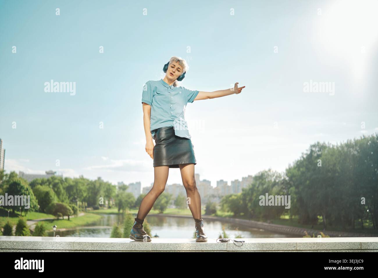 Femme se tient sur le rebord près de la rivière portant chemise bleue et jupe noire. Elle danse librement avec des écouteurs. Les arbres verts et les bâtiments sont visibles à l'arrière Banque D'Images